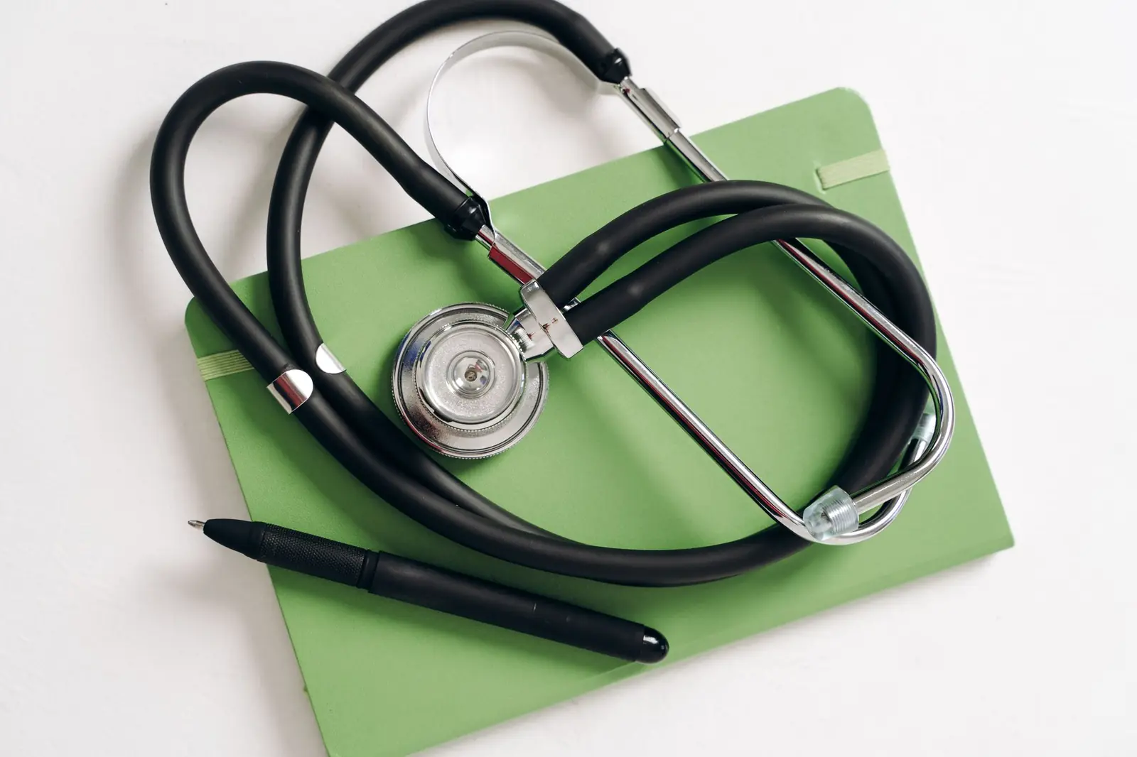 A stethoscope and black pen resting on a green notebook on a clean white desk.