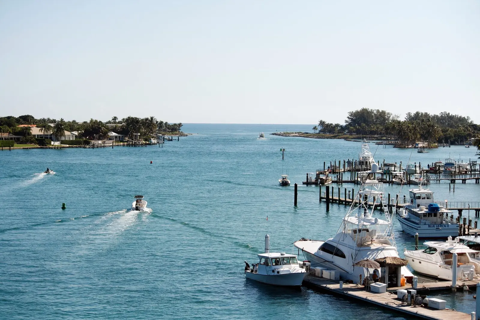 Boats heading out through Jupiter Inlet past docked fishing yachts and palm-lined shores on a clear day.