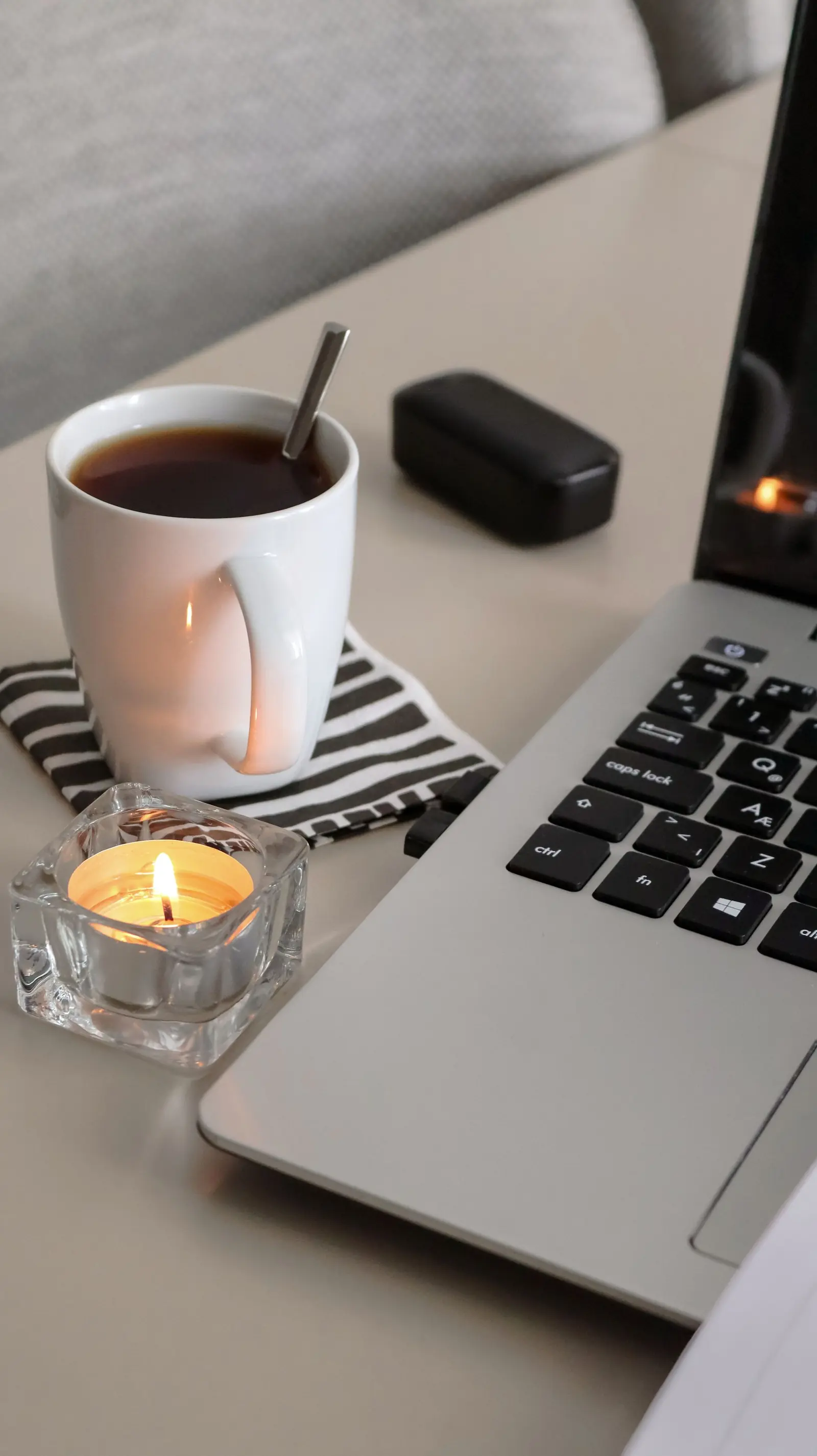 A laptop beside a cup of tea and a lit candle on a quiet desk at home.
